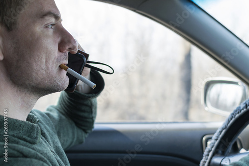 A young man rides in a car with a cigarette in his mouth and a dissatisfied face talking on the phone.