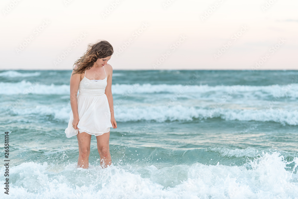 Young woman standing in white dress on beach sunset in Florida panhandle with wind ocean waves looking down Young woman standing in white dress on beach sunset in Florida panhandle with wind ocean waves looking down