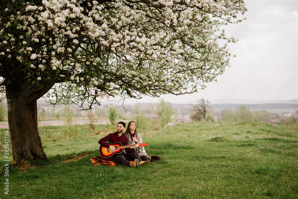 Young couple sitting under the big tree and man playing on the guitar