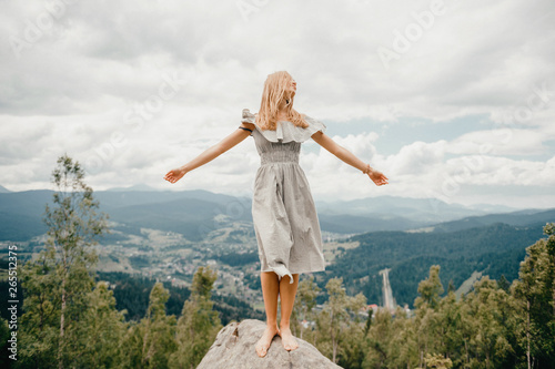 Young beautiful barefoot blonde girl with long hair in summer dress standing on top of conquered mountain at stone and enjoying fabulous landscape scenic view with mountains and village in valley
