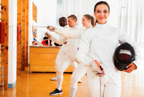 Smiling sporty young woman in uniform standing at fencing workout