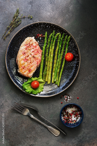 Grilled chicken fillet in a plate with asparagus, tomato and spices on a dark background. Overhead view,flat lay.
