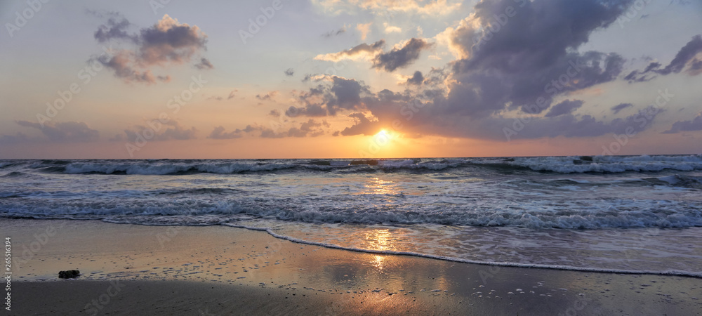 Breathtaking early spring sunset at the beach in Netanya, Israel!  Bright orange sun is setting down over the horizon, beautiful  fluffy clouds hanging over the foamy, wavy sea. 