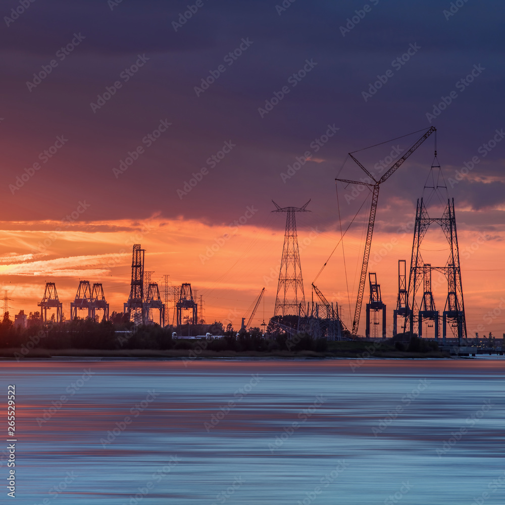 Riverbank with silhouettes of container terminal cranes during an orange colored sunset, Port of Antwerp, Belgium.
