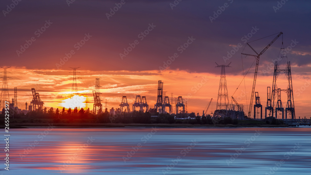 Riverbank with silhouettes of container terminal cranes during an orange colored sunset, Port of Antwerp, Belgium.