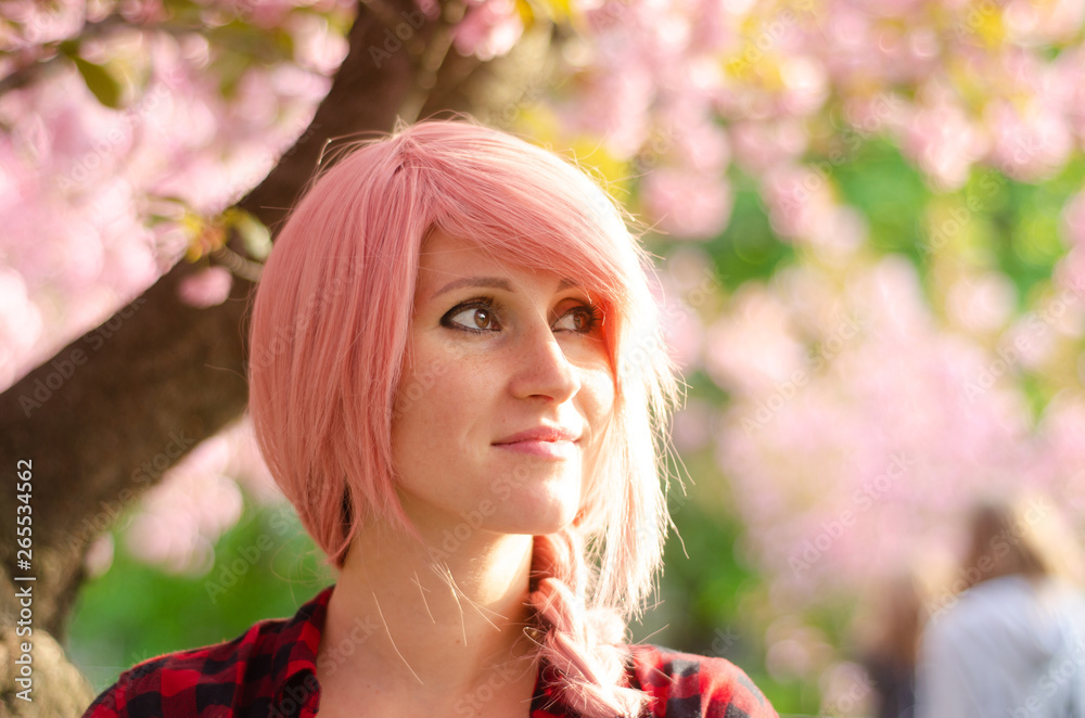 Fototapeta premium Young hipster women with pink hair is enjoying time outdoors. Happy beautiful face. Natural look. Green nature spring background with pink sakura flowers