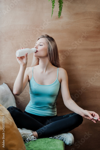 Young beautiful athletic woman blonde in sportswear sitting by the window of a cafe against the background of a wooden wall.