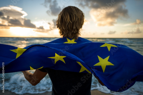 Man with blond hair holding a fluttering EU European Union flag on a sunrise beach in front of stormy turbulent Channel seas