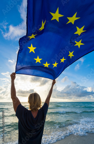 Man with blond hair holding EU European Union flag flying in the wind on a sunrise Mediterranean Beach