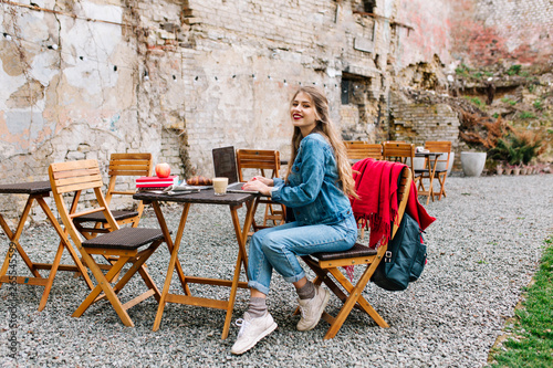 Charming blonde female freelancer using laptop for remote job during breakfast in the outdoor cafe. Portrait of young woman using computer in the park. Distant job, smiling girl, workplace