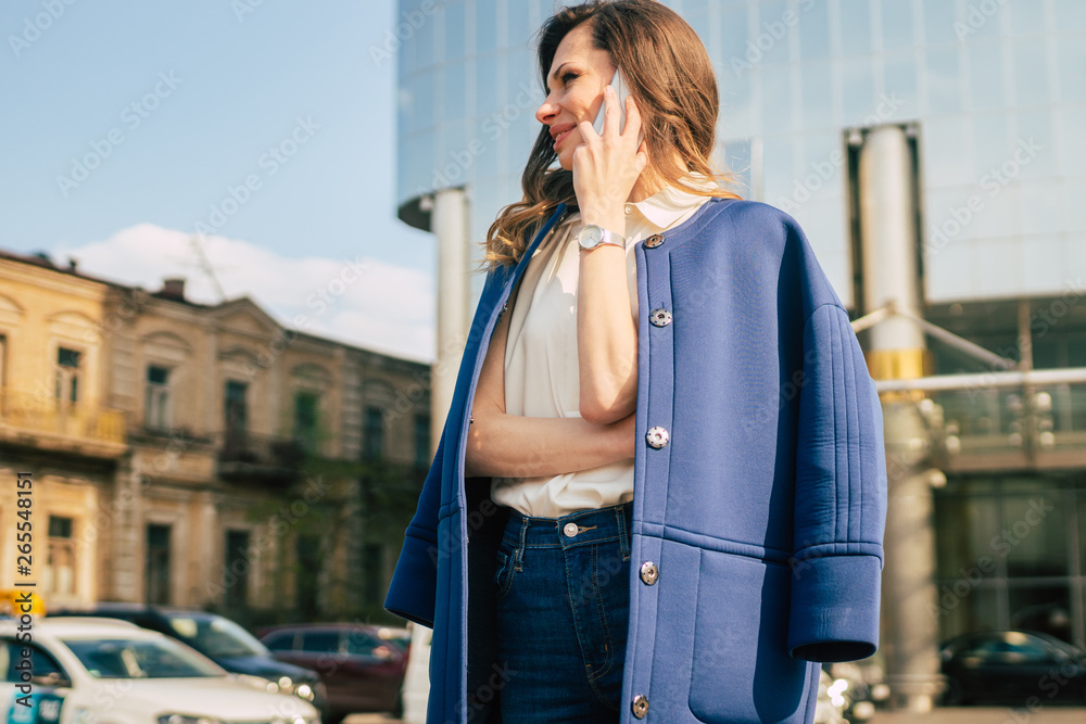 Fototapeta premium Caucasian business woman speaking by phone. Waist up portrait of a successful European business woman woman, talking on the phone, standing on glass background, modern office building. Sunny weather