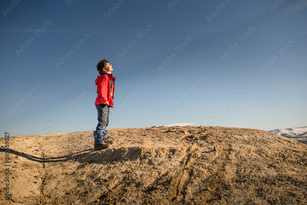 Young boy in red jacket facing sun on top of mountain with blue sky