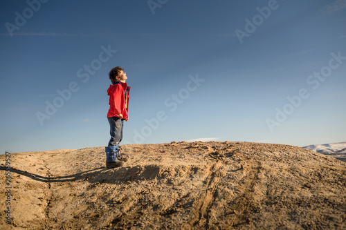 Young boy in red jacket facing sun on top of mountain with blue sky