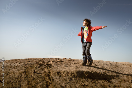 Young boy in red jacket on mountain facing sun with arms outstretched and hair blowing