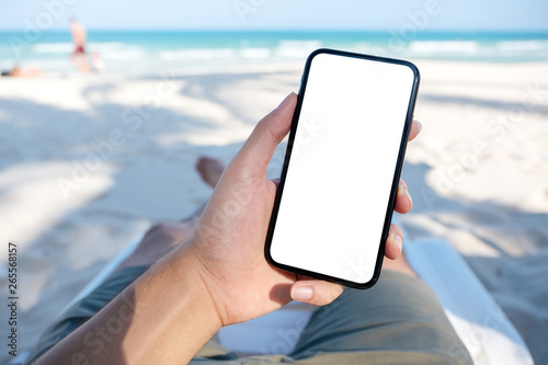 Fototapeta Naklejka Na Ścianę i Meble -  Mockup image of a man's hand holding white mobile phone with blank desktop screen while laying down on beach chair