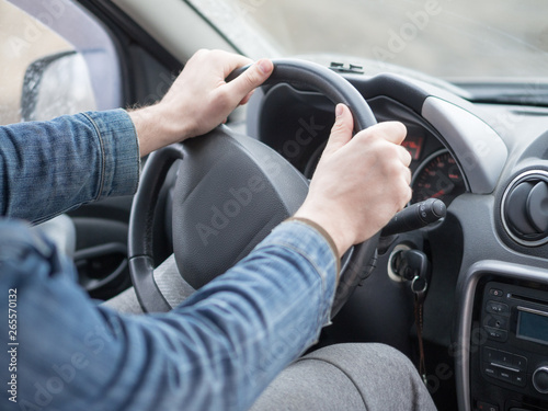 Human arms in denim sleeves on steering wheel, inside cab view, close up, selective focus. Ordinary man driving a car.