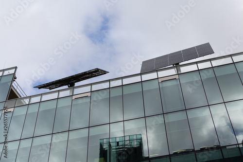 Large rotating solar panels on the roof of a large glass commercial office building against a cloudy sky.