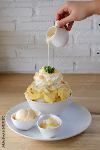 Pouring sweetened condensed milk on durian bingsu served with vanilla ice cream and sticky rice on wooden table