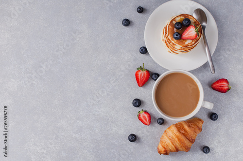 pancakes with berries and a cup of fresh coffee on the right side, croissant, grey background