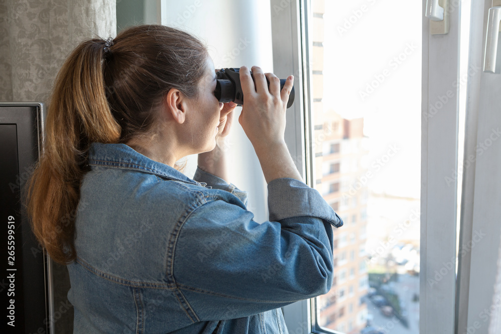 Adult woman spying out the window looking through binoculars, a domestic room
