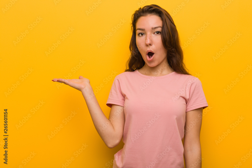 Young european woman isolated over yellow background impressed holding copy space on palm. Young european woman isolated over yellow background impressed holding copy space on palm.