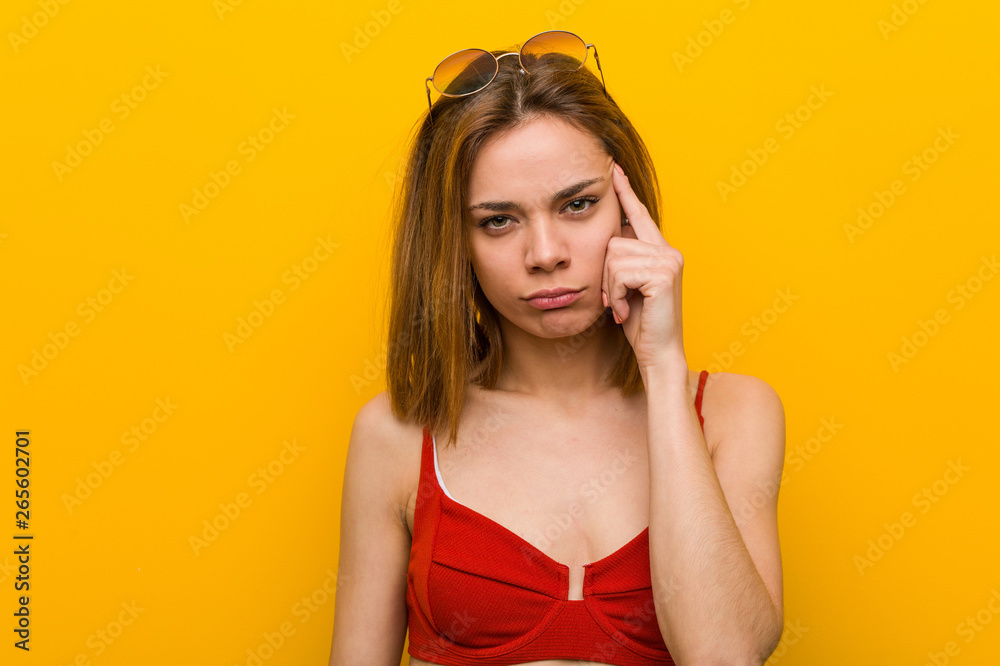 Young caucasian woman wearing bikini and sunglasses pointing her temple with finger, thinking, focused on a task. Young caucasian woman wearing bikini and sunglasses pointing her temple with finger, thinking, focused on a task.