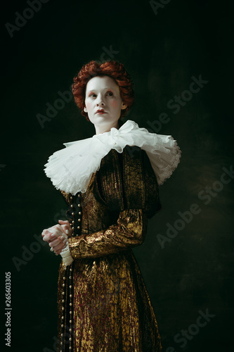 No fear for future. Medieval redhead young woman in golden vintage clothing as a duchess standing crossing hands on dark green background. Concept of comparison of eras, modernity and renaissance.