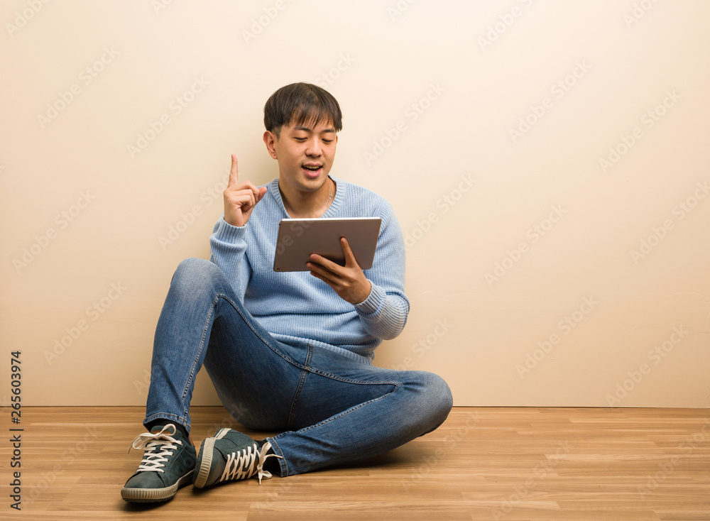 Young chinese man sitting using his tablet surprised pointing up to show something Young chinese man sitting using his tablet surprised pointing up to show something