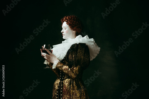 One part of me. Medieval redhead young woman in golden vintage clothing as a duchess holding a glass with red wine on dark green background. Concept of comparison of eras, modernity and renaissance. photo