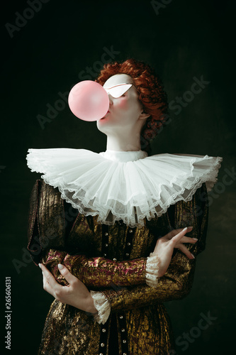 Medieval redhead young woman in golden vintage clothing as a duchess with red sunglasses blowing a bubblegum on dark green background. Concept of comparison of eras, modernity and renaissance. photo