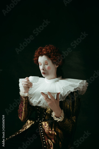 Too hot and strong. Medieval redhead young woman in golden vintage clothing as a duchess drinking a cup of coffee on dark green background. Concept of comparison of eras, modernity and renaissance.