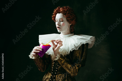Taste of future. Medieval redhead young woman in golden vintage clothing as a duchess eating a fried potato on dark green background. Concept of comparison of eras, modernity and renaissance.