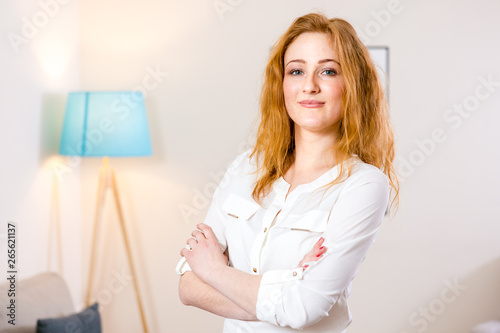 young Caucasian girl standing in the office and folded her arms across her chest. Charming young girl works inside in bright office, coworking room. The girl has blond red hair and white shirt © Elizaveta