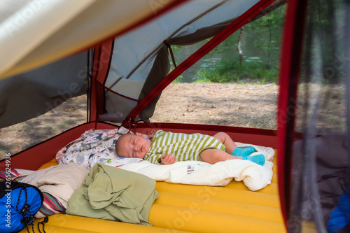 Little boy is sleeping on his back in green striped clothing inside a tent not far from summer lake