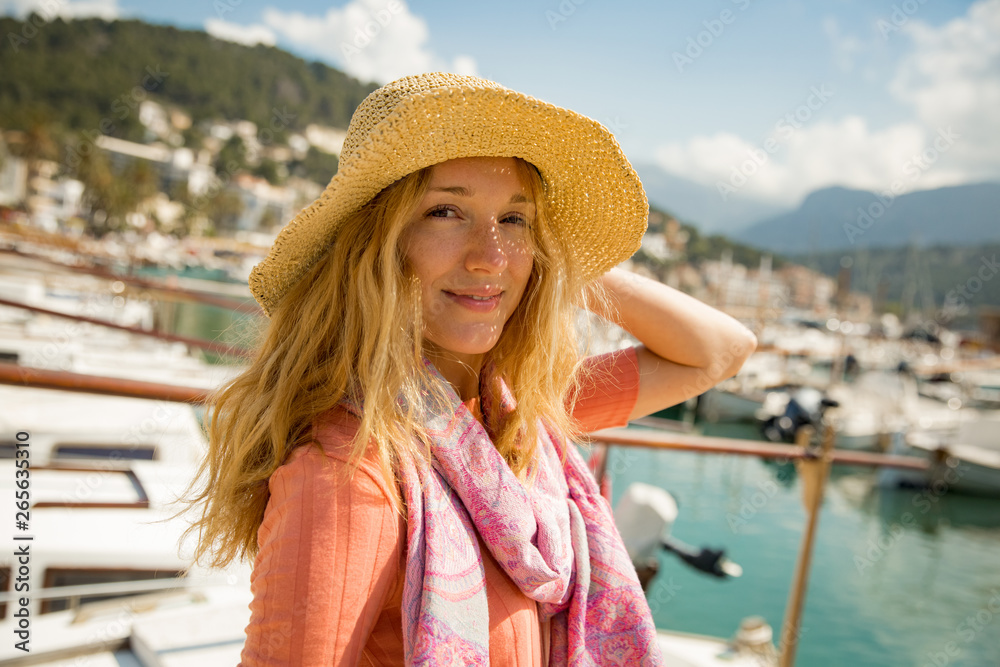 Portrait of young woman with light curly hair, holding straw hat with hand, enjoying sun breeze, smiling. Sunny harbor, boats and yachts, green mountains on background. Enjoying life, happy traveling,