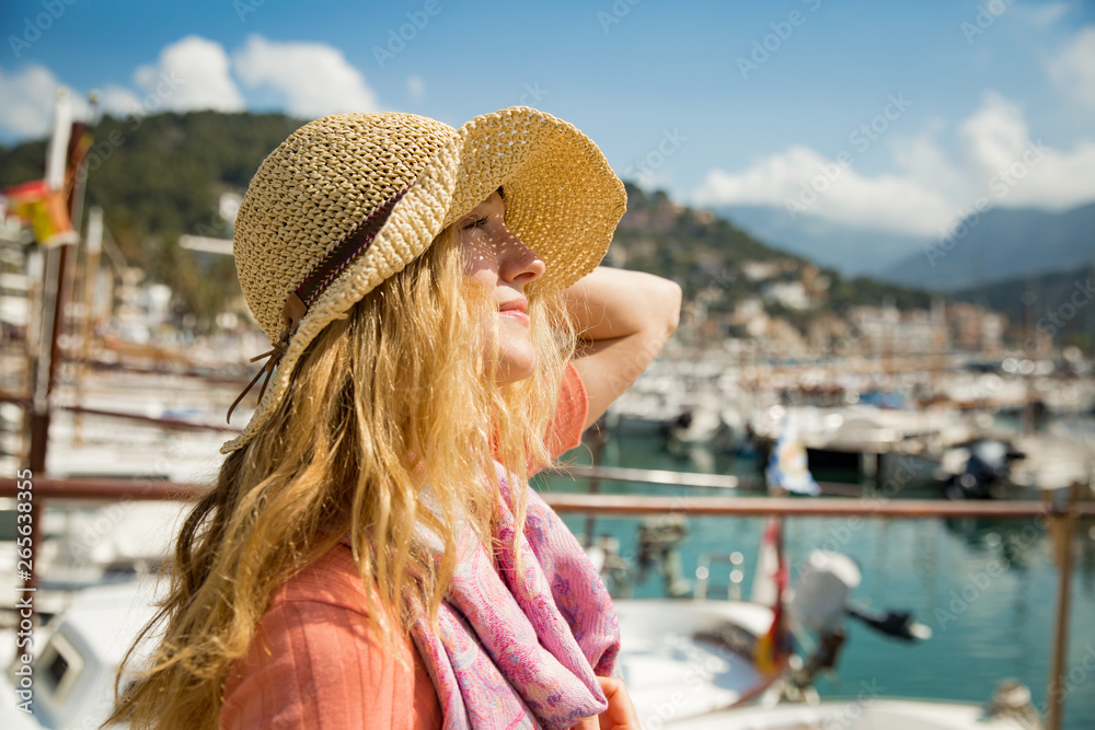 Portrait of young woman with light curly hair, holding straw hat with hand, enjoying sun breeze, smiling. Sunny harbor, boats and yachts, green mountains on background. Enjoying life, happy traveling,