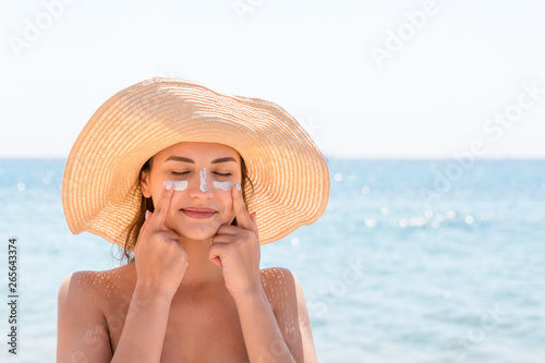 Beautiful young woman in hat is applying sunblock under her eyes and on her nose like Indian. Sun protection concept photo