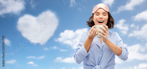 valentine's day, people and bedtime concept - happy young woman in pajama and eye sleeping mask drinking coffee from mug over background