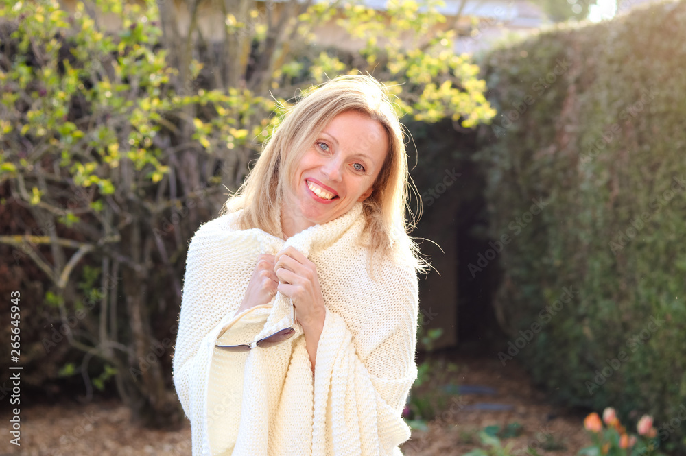Happy attractive joyful blond girl in ivory knitted plaid smiling and looking at camera outdoors in garden at fresh sunny evening. Lifestyle Happy attractive joyful blond girl in ivory knitted plaid smiling and looking at camera outdoors in garden at fresh sunny evening. Lifestyle