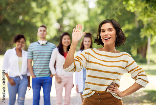 gesture and people concept - happy smiling young woman in striped pullover waving hand over group of friends in summer park background