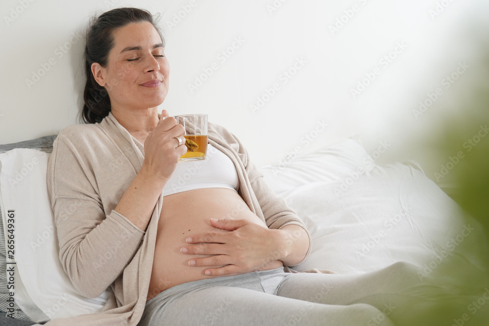Pregnant woman laying on bed and drinking infusion Pregnant woman laying on bed and drinking infusion