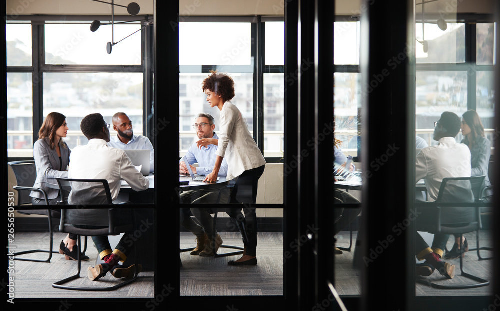 Millennial black businesswoman stands addressing colleagues at a meeting, seen through glass wall