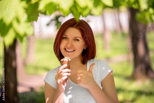 Portrait of a smiling woman standing under trees in park or forest with big ice-cream and showing thumb up. Enjoying the moment and summer/spring relax concept