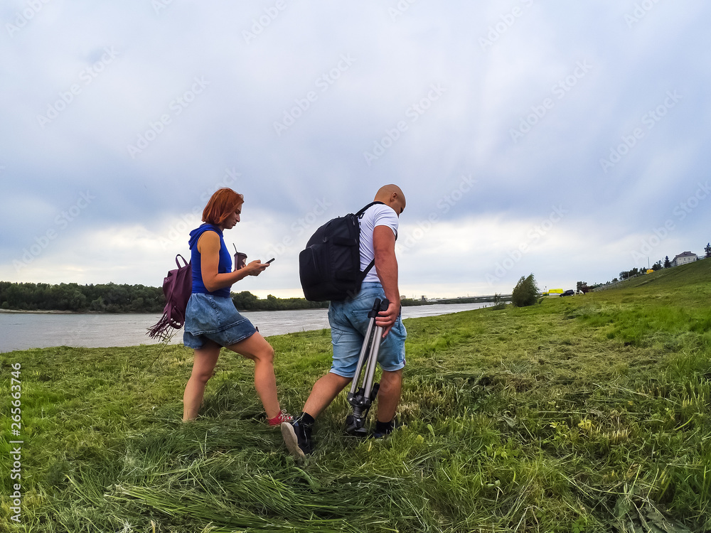 The guy and the girl in shorts and T-shirts go on high green grass on the river bank with backpacks. A woman has coffee and a telephone in her hands, a man has a tripod for a camera. Walk and sports.
