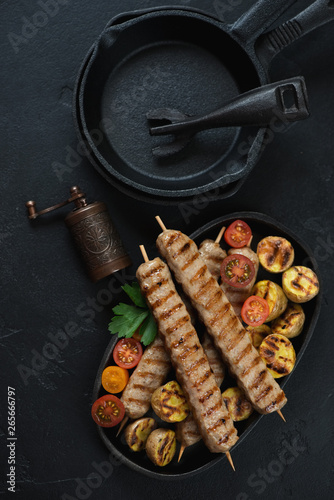 Barbecued chicken skewers and cast-iron frying pans over black stone background, view from above, vertical shot