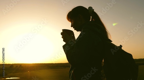 silhouette woman traveler, stands on top of hill drinking coffee in glass from thermos. tourist girl drinks hot tea and looks at sun. Adventurer rest after reaching goal. freedom concept.