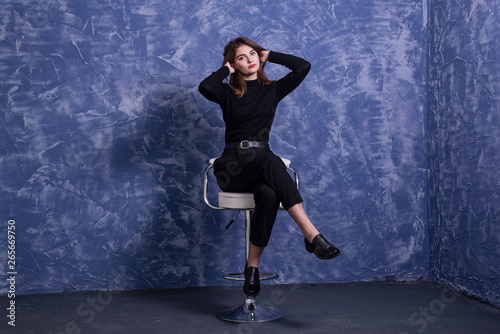 A young woman is sitting on a bar stool against the background of a blue wall, free space.