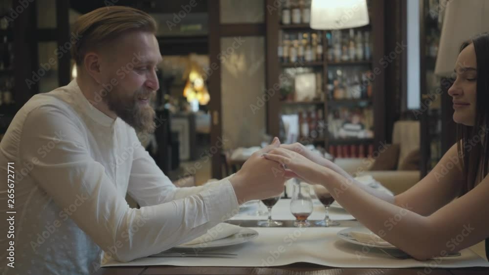 Glamour brunette woman and a handsome bearded blond man sitting at the table in front of each other. The man telling good words to his girlfriend then kisses her hand. The cute couple has a date Glamour brunette woman and a handsome bearded blond man sitting at the table in front of each other. The man telling good words to his girlfriend then kisses her hand. The cute couple has a date