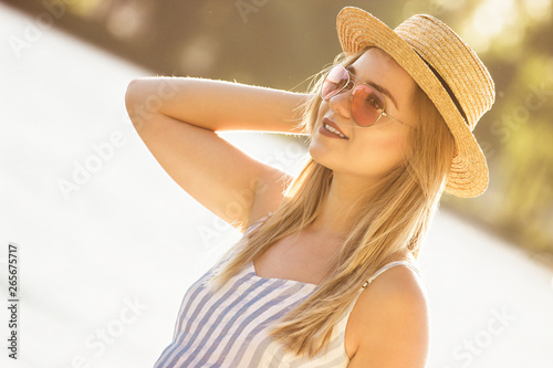 Portrait of tender young woman in straw hat and sunglasses standing at sunny riverside. Beautiful stylish girl in stripped overalls posing at beach on summer sunset. Girl in summer dress on beach