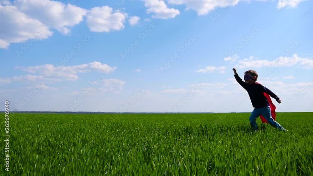 A little boy in a superhero costume turns around running across a green field holding his hand forward A little boy in a superhero costume turns around running across a green field holding his hand forward